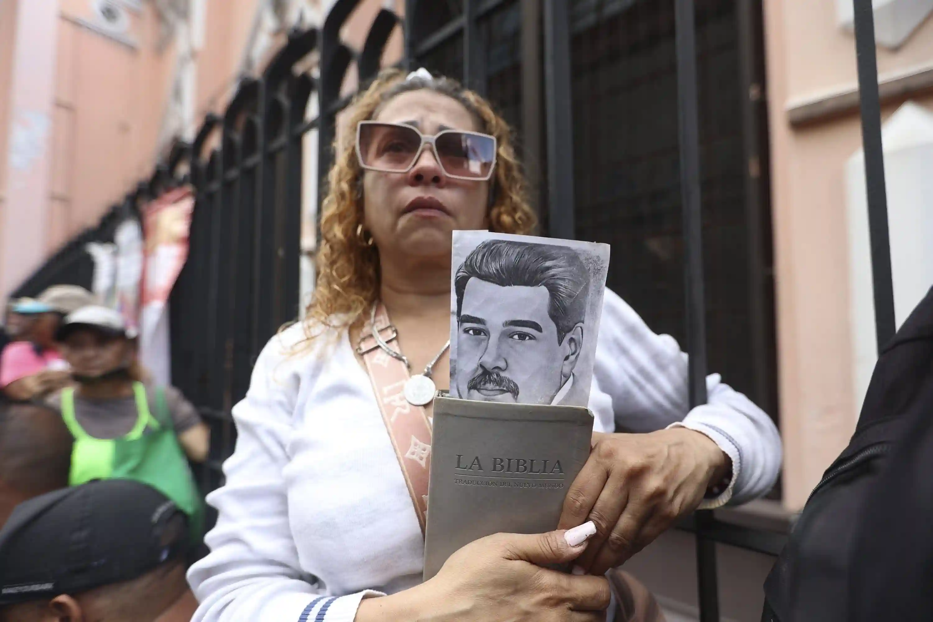 Woman holds a Bible and a portrait while standing outside a gated building during a public gathering