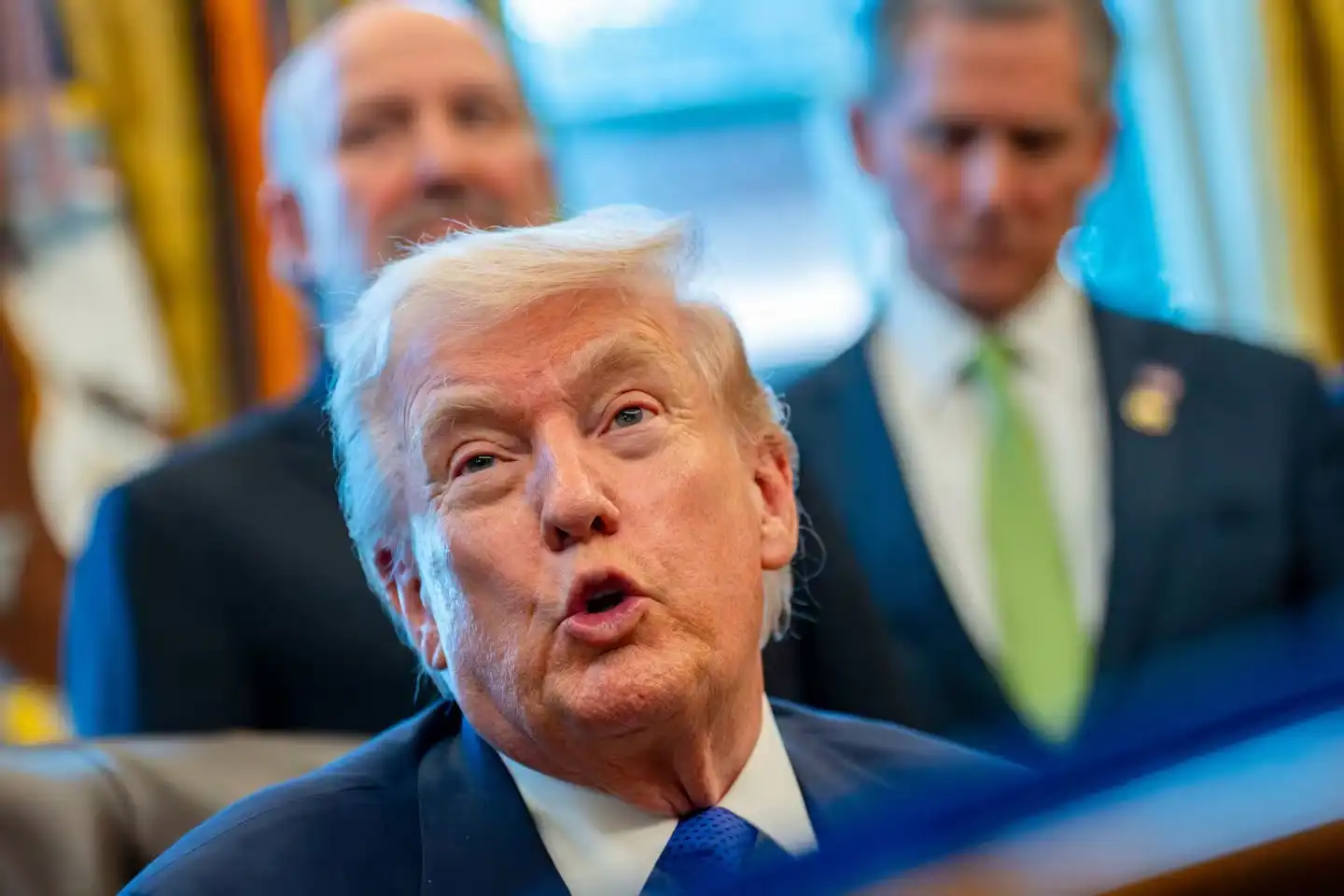 A close-up view of President Donald Trump speaking in the Oval Office, with individuals in the background during a meeting.