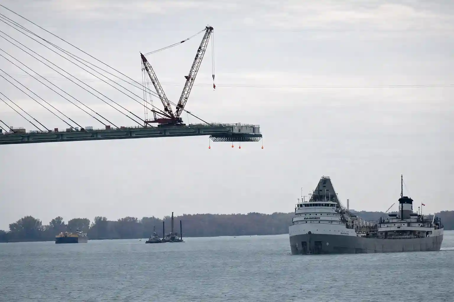 Construction cranes working on the nearly completed Gordie Howe International Bridge over the Detroit River, with a cargo ship passing below